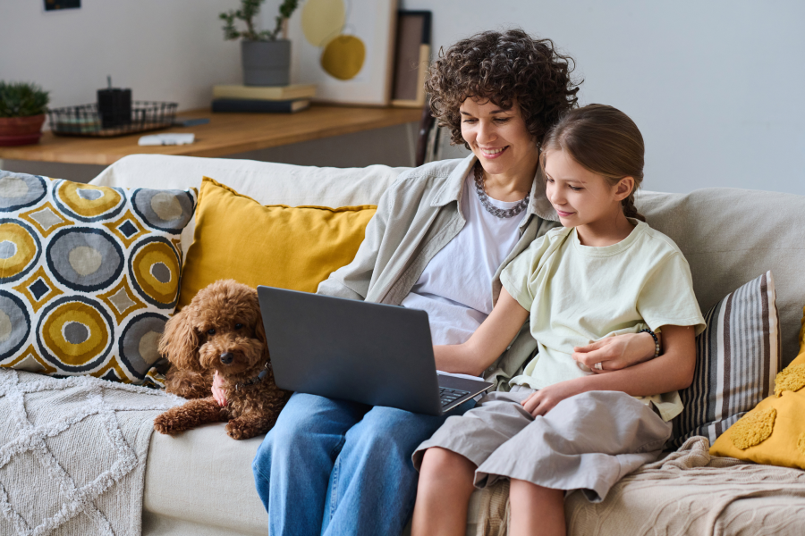 A smiling mother and young daughter sitting together on a comfortable couch using a laptop for home research.